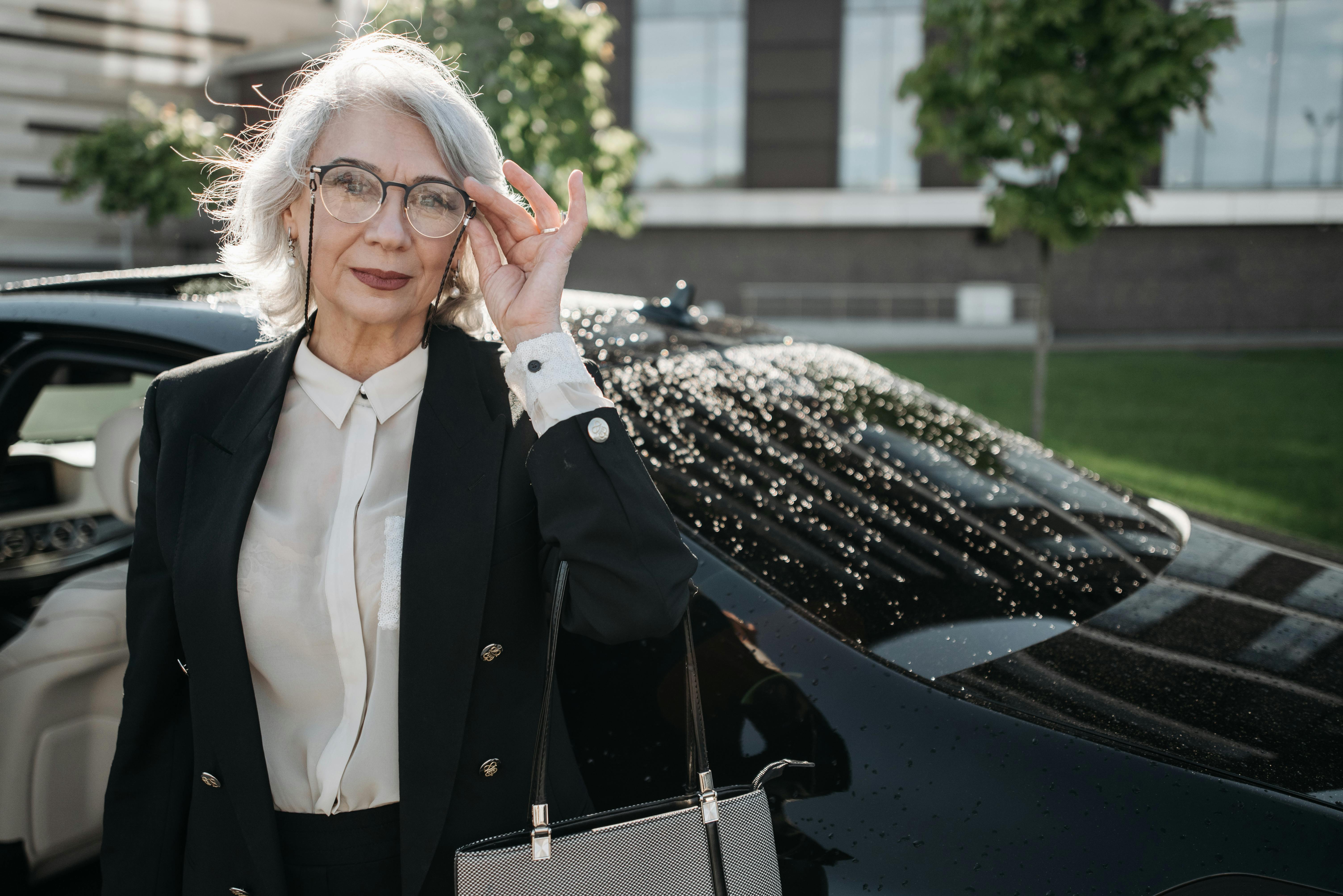 Stylish senior businesswoman with grey hair and eyeglasses standing by a black car outdoors.